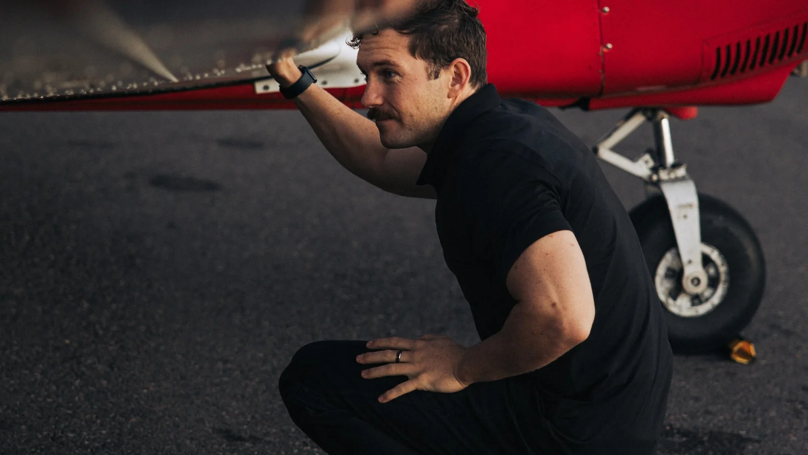 A man crouches under the wing of a small red and white aircraft, checking the underside beside the main landing gear.
